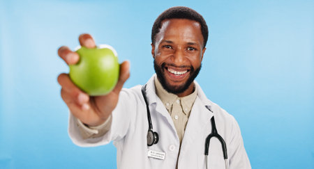 Healthy, doctor and black man with apple in portrait for nutrition, healthcare diet or food choice. Studio, blue background or happy African nutritionist with fruit for fibre, snack and vitamin cの写真素材