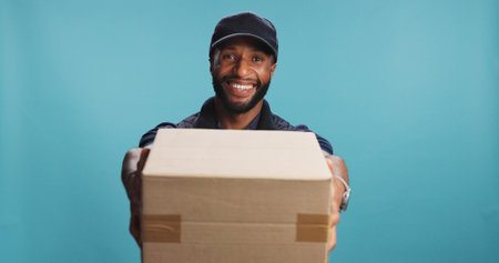 Portrait, delivery man and offer box for shipping, logistics or supply chain. African courier, face and giving package for distribution service, ecommerce or smile for order on blue studio backgroundの写真素材