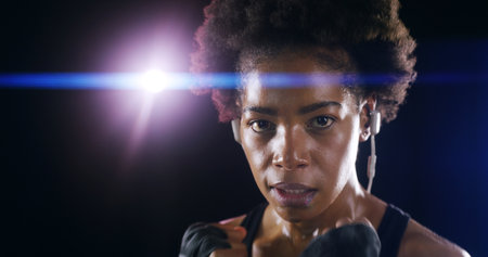 Woman, boxer fighter and studio for workout, sweating and lens flare on black background. African person, performance training and strong power with martial arts, portrait and sport song on earphonesの写真素材