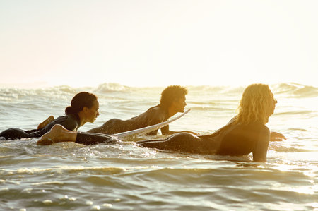 Sunset, travel and friends surfing at the beach while on a summer vacation, adventure or weekend trip. Watersports and group of people practicing to surf in the ocean at twilight on tropical holiday.の写真素材