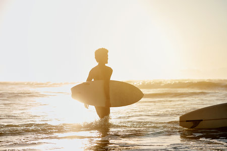 Back, surf and sunset with a sports man in the ocean during summer for hobby, leisure or recreation. Surfing, sea and nature with a male athlete or surfer walking into the water for health or fitnessの写真素材