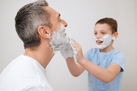 Shaving, father and child with cream in studio for grooming, skincare lesson and product application. Learning, mature man and boy kid with foam for cleaning face and hair removal on white backgroundの写真素材