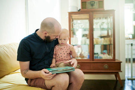 Dad, baby and reading book in a family home living room for fun. Man or dad with toddler kid on couch for quality time, love and care or learning, talking and together for story or developmentの写真素材