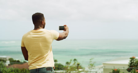 Travel, photography and man at ocean with phone, memory and tropical vacation at hotel from back. Holiday blog, live stream and influencer at beach with smartphone for online review for island resortの写真素材