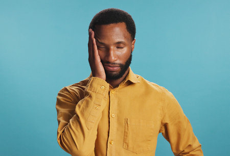 Stress, tired and black man with headache in studio, tension or pressure on blue background. African person, burnout or model with migraine, illness or frustrated with brain fog, pain and fatigueの写真素材
