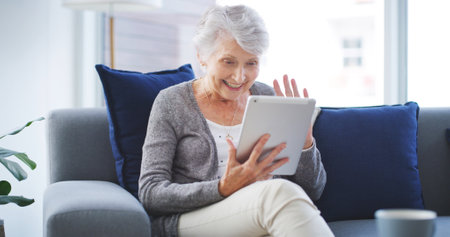 Video call, greeting and senior woman with tablet on sofa in living room for telehealth communication. Happy, waving and elderly person on virtual medical consultation on digital technology in home.の写真素材