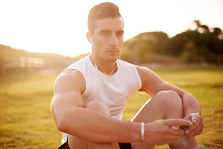 Man, fitness portrait with headphones on grass for exercise, break and listening to music in nature. Serious, athlete and sunrise with tech for outdoor workout playlist, training and run in morningの写真素材