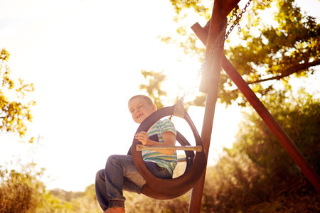 Sky, swing and portrait of boy in park with trees, outdoor adventure and child development in nature. Play, smile and kid in playground for growth, fun games or summer childhood on weekend from belowの写真素材