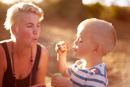 Happy, blowing bubbles and child in park with mom for fun, childhood and bonding together outdoors. Family, sunlight and woman with young boy with soap wand, toys and activity for playing in natureの写真素材