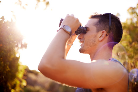 Man, happy and binoculars in nature for bird watching, explore or enjoy view in Italy. Person, profile or outdoor with magnifying lens for adventure, sightseeing or travel in countryside for wellnessの写真素材