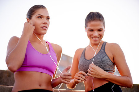City, women and smile with earphones for fitness, workout playlist and listening to audio outdoor. Happy people, sisters and break for training with music, wellness or podcast for exercise motivationの写真素材