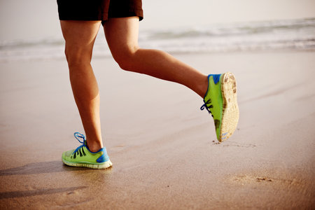 Man, sand and legs running in nature for fitness, outdoor cardio and sports workout for exercise. Male person, sneakers and athlete for training by water, ocean and performance on tropical holidayの写真素材