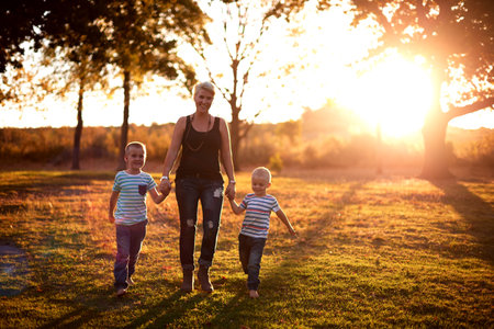 Happy mother, walking or holding hands with children in nature for bonding holiday or sunset at outdoor park. Mom, kid or siblings on grass field with sunlight or trees for playful day in countrysideの写真素材