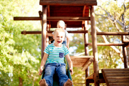 Excited, playing and portrait of children on slide in park for physical skills, confidence or fun. Exploring, nature and boy kid son playground equipment for adventure, hobby or cognitive developmentの写真素材