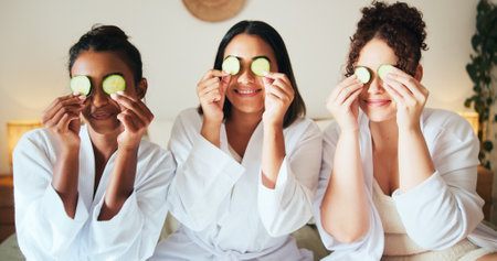 Cucumber, skincare and smile with friends in bedroom for beauty, spa day and dermatology. Facial, natural cosmetics treatment and hotel resort with women for self care, luxury and relax togetherの写真素材