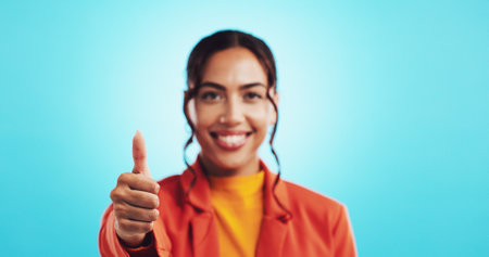 Woman, thumbs up and winner emoji in studio, promotion and agreement portrait on blue background. Motivation, success gesture and like symbol for thank you, great and feedback sign for achievementの写真素材