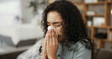 Woman, blowing nose and home for respiratory influenza, allergies and sinus disease or infection. Couch, viral sickness and toilet paper to remove bacteria in lounge, fatigue and tissue for hayfeverの写真素材