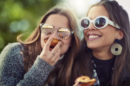 Friends, women and eating fruit in park with sunglasses for nutrition, healthy snack and bonding in nature. Happy people, together and fresh peach for relax, summer holiday and organic diet outdoorの写真素材