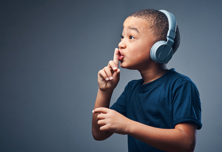 Black child, finger and lips for secret in studio with headphones for announcement, news and gossip. Boy, listening and music for privacy sign, confidential story and silence noise on gray backgroundの写真素材