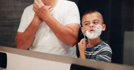 Shaving, learning and child with father in bathroom for grooming, hygiene and morning routine. Cream, razor and dad teaching boy kid with facial hair removal by mirror for skincare in home together.の写真素材