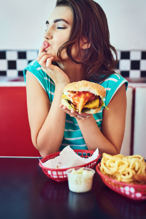 Girl enjoying and eating a burger in restaurant for lunch meal craving, unhealthy snack and hungry at table. Customer, person or fast food brunch in retro diner for hamburger sandwich or diningの写真素材
