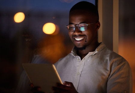 Office, night and business black man on tablet for research project, online website and planning. Happy, creative and person on digital tech working late for networking, internet news and reportの写真素材