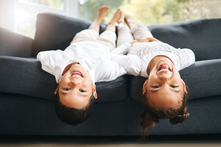 Portrait, upside down and happy kids on couch laughing at funny joke with joy or games in family home. Girl, silly boy and playful children siblings bonding on sofa playing together in house to relaxの写真素材