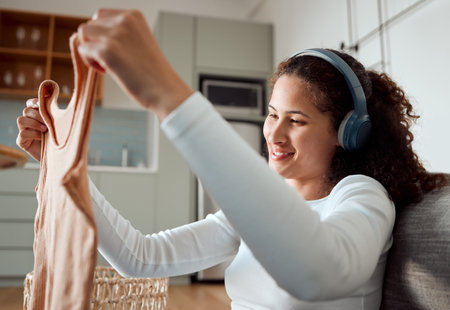 Woman, headphones and tidy laundry in home, housework and listening to music in apartment. Female person, fold washing and packing clothes for textile maintenance, cleaner and hearing sound or audioの写真素材
