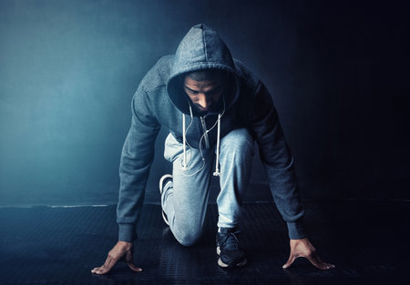 Running, start and man in studio with challenge, health and wellness in hoodie at gym workout. Fitness, ready and confident runner with marathon training, commitment and exercise on dark backgroundの写真素材