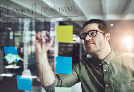 Businessman, writing or reminder with glass board for brainstorming, agenda or schedule planning at office. Man, creative or employee with sticky note for startup project, tasks or ideas at workplaceの写真素材