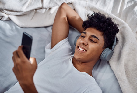 Phone, happy man and listening to music in bed, audio and sound to relax in home. Mobile, bedroom and person on headphones for streaming radio, hip hop podcast and reading message on social mediaの写真素材