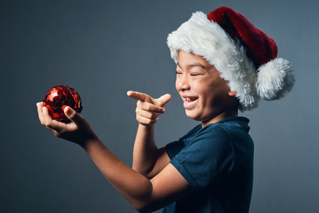 Happy boy, christmas and pointing with red ball for decoration, festive or holiday on a gray studio background. Young, male person or laughing with santa hat or ornament for December celebrationの写真素材