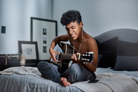 Guitar, bedroom and man with earphones in home playing music for learning chords, tuning or songwriting. Artist, acoustic instrument and male person relaxing on with strings for creativity at house.の写真素材