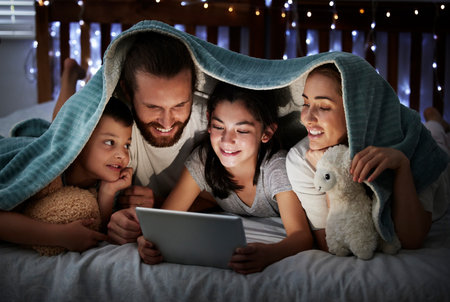 Happy caucasian family with two children using digital tablet lying under blanket in the dark at night with their faces illuminated by device screen light. Family of four reading online story or watcの写真素材