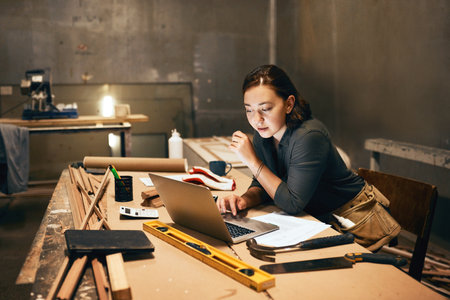 Woman, wood and laptop in workshop for planning, design process and research at night. Carpenter, tech and connectivity in studio for material order, furniture creativity or small business managementの写真素材