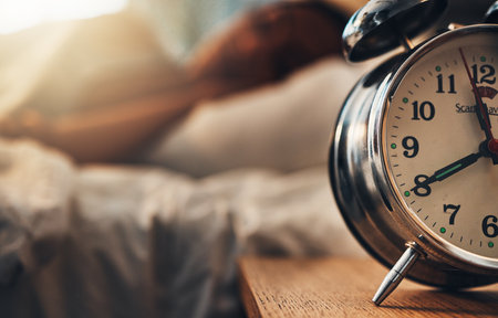 Alarm clock, table and person sleeping, relax and resting in bedroom for energy or lazy at home. Time, bed and oversleeping with fatigue, morning and tired on comfortable mattress for peace or calmの写真素材