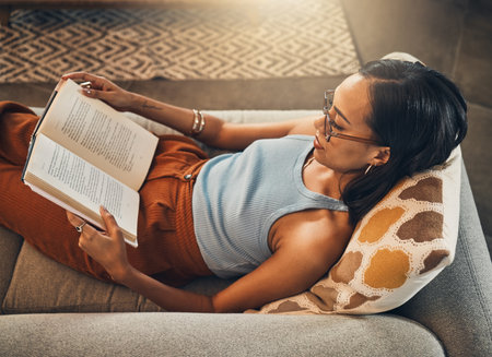 Book, reading and relax with woman on sofa in living room of home for knowledge or wellness from above. Hobby, literature and storytelling with person in apartment on weekend for break or recoveryの写真素材