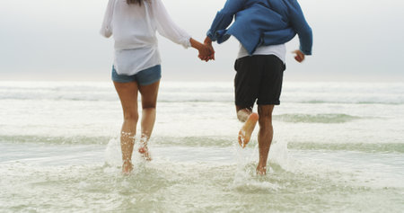 Couple, holding hands and running in water at beach for travel holiday, freedom or support in relationship. Back, legs of people and romantic date by ocean with adventure, trust or honeymoon vacationの写真素材