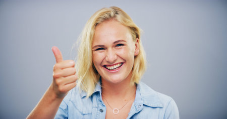 Happy woman, portrait and feedback with thumbs up for good reaction on a gray studio background....
