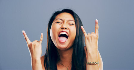 Rocker, hand and crazy with portrait of Asian woman in studio for metal music, punk identity and crazy. Devil horn sign, edgy and attitude with person on background for energy, emoji and expressionの写真素材