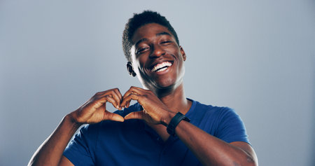 Heart hands, portrait and smile with happy black man in studio for affection, love or romance. Emoji, flirt and social media with African person on gray background space for health or wellnessの写真素材