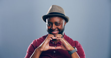 Heart hands, portrait and smile with African man in studio for affection, love or romance. Emoji, hat and social media with happy black person on gray background mockup for health or wellness gestureの写真素材