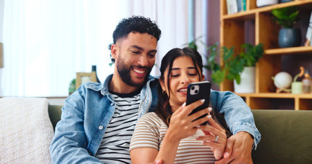 Couple, smartphone and social media in living room with hug, bonding and funny story in home. Woman, man and phone on sofa for storytelling, comedy and celebrity news on timeline with internet gossipの写真素材
