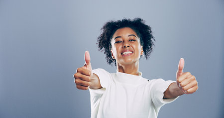 Portrait, thumbs up and woman with agreement, like and confidence on gray studio background. Face, happy person and model with hand gesture, feedback and icon with symbol, perfect and like with emojiの写真素材