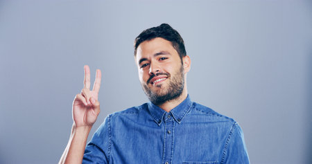 Portrait, creative and happy man with peace sign in studio with positive attitude, confidence or facial expression. Grey background, casual and proud male model for cool gesture, fashion or v symbolの写真素材