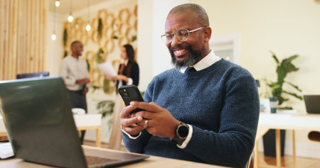 Phone, creative and business man in office for email, notification or magazine editor typing review. Startup, mobile and happy mature manager online, reading article and check publication on laptopの写真素材