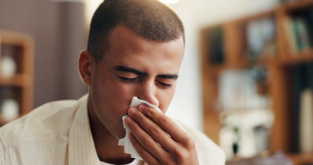 Sick man, flu and blowing nose with tissue for influenza or cold with allergy, sinus or hayfever at home. Male person, germs and bacteria with sneeze, mucus or cough for viral infection or virusの写真素材
