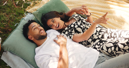 Outdoor, pointing and couple on date, picnic and smile for conversation in nature, embrace and relationship. Happy, man and bonding with woman on anniversary, people and cloud watching for romanceの写真素材