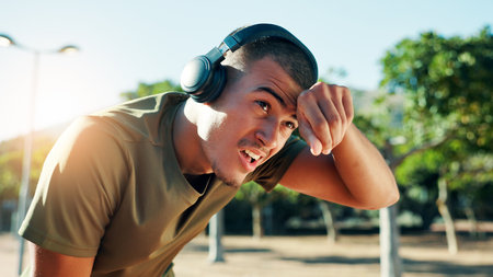 Tired, man and breathing with headphones for fitness at park with running fatigue, workout music or break. Athlete, sweating and exhausted in nature with exercise podcast, resting or cardio challengeの写真素材