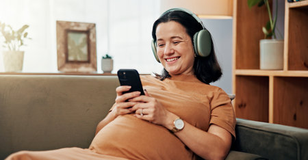 Pregnant woman, phone and headphones on sofa for smile, texting or search for music on mobile app in home. Person, smartphone and relax with streaming, subscription or video on social media in houseの写真素材
